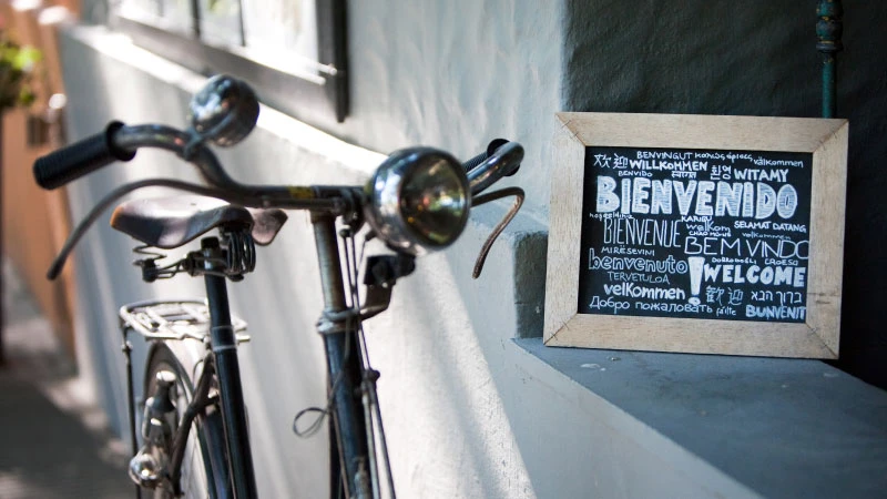 Bicicleta estacionada en la entrada de un bar con un cartel de bienvenida en diferentes lenguajes